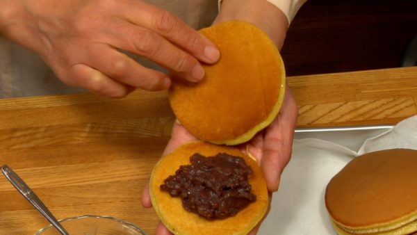 Let's make regular Dorayaki. Take the pancakes and hold, lightly browned side facing up. Spread spoonfuls of anko on the middle of the pancake. Place another pancake on top and press around the edges to shape. Serve it on a plate.