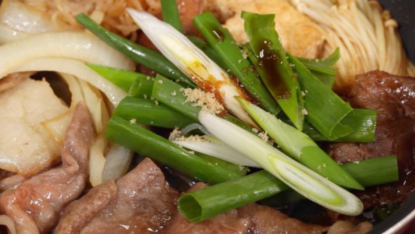 We will show you how to add the ingredients to the sukiyaki. For example, adding the spring onion leaves. When you place new ingredients into the sukiyaki, add the seasonings for them. Sprinkle on the sugar. And add the soy sauce. Combine the seasonings, and the spring onion leaves are ready.