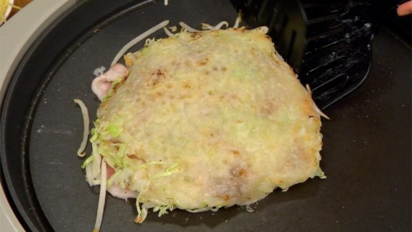 Gira l'okonomiyaki con delle spatole apposite. Raduna gli ingredienti che possono essersi sparsi per la piastra e ricomponi il tutto.