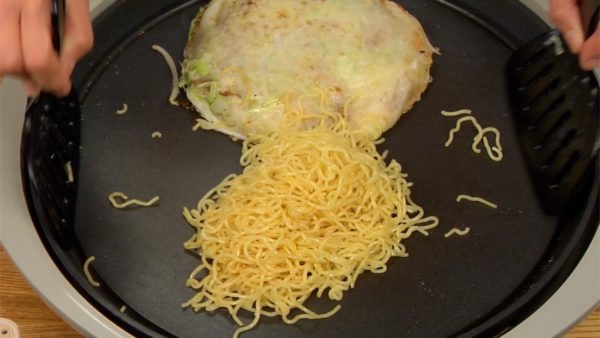 Añadir la salsa okonomiyaki y continuar friendo, distribuyendo la salsa uniformemente. Darle forma de círculo a los fideos y dorar el otro lado.