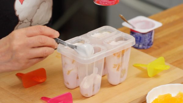 Nous allons utiliser des moules à glaces. Versez un peu de yaourt à la myrtille dans les moules. Ajoutez 2 morceaux de kiwi. Ensuite, ajoutez dessus un peu de yaourt à la fraise. Ajoutez 2 morceaux d'orange. 
