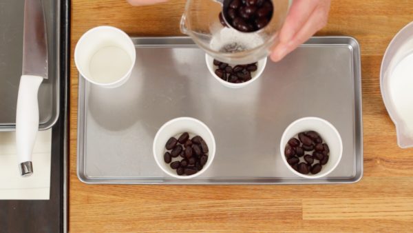 Kami akan membuat es loli susu azuki dengan menggunakan gelas kertas kecil. Masukkan 1 sendok makan amanatto, sejenis permen kacang azuki pada setiap gelas.