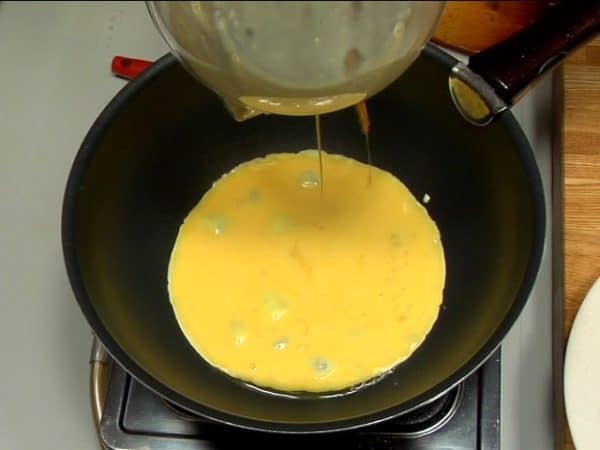 Vamos fazer o Omurice. Adicione azeite de oliva à uma frigideira quente e gire-a para que todo o fundo fique coberto com o azeite. Para ver se a temperatura está adequada, coloque um pouquinho de ovo na frigideira. Então adicione toda a mistura de ovo de uma vez só.