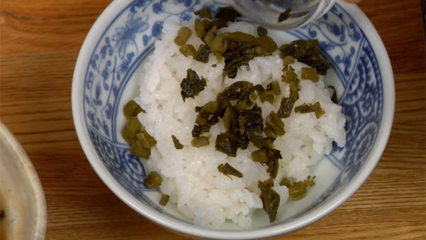 Tiếp theo, chúng tôi sẽ làm một loại Ochazuke khác. Để nhẹ cơm nóng vào bát ăn cơm. Rắc takana-zuke đã thái lên. Để lá tía tô đã thái, cá mòi trắng (shirasu) và umeboshi vào trung tâm cơm. Rắc hạt mè rang lên.
