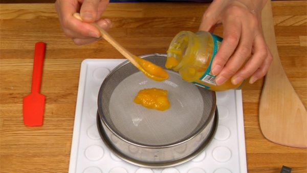 Let’s purée the mango jam. Spoon the jam onto a flat, or uragoshi strainer. Press the jam with a wooden paddle and continue scraping toward you. Removing the lumps from the jam will prevent the mochi from breaking later.