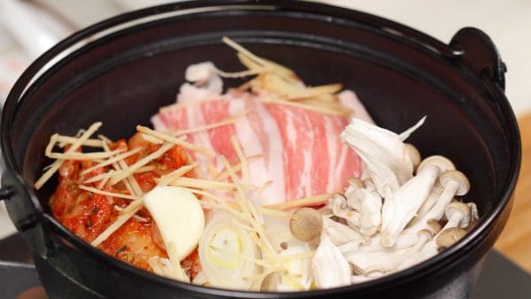 Thêm các lát thịt lợn (heo), kimchi (rau ngâm kiểu Hàn Quốc, hành boa rô đã thái và nấm đùi gà, sau đó là tép tỏi và gừng thái sợi.