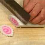 Affettare sottilmente il narutomaki, un tipo di impasto di pesce e amido, cotto a vapore, caratterizzato da un disegno a spirale rosa al centro e una forma cilindrica.