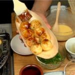 Place 8 takoyaki pieces onto a ship shaped wooden plate and coat them in the okonomiyaki sauce with a pastry brush.