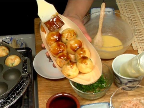Để 8 miếng takoyaki lên đĩa gỗ hình con thuyền và phủ chúng bằng sốt okonomiyaki bằng chổi quét bánh.