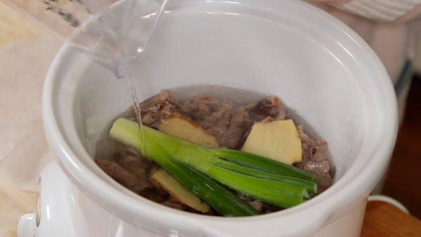 Now, place the gyusuji into a slow cooker. Add the ginger root slices, green part of the long green onion, sake and water.