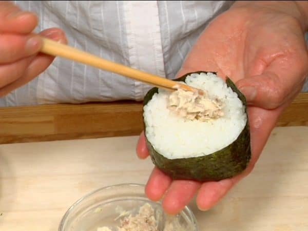 Das Onigiri mit dem langen Nori-Streifen umwickeln. Oben eine kleine Vertiefung eindrücken und mit der restlichen Füllung dekorieren.