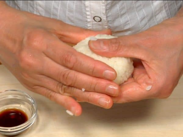 And now we’ll make a delicious soy sauce onigiri! Wet your hands with the soy sauce and firmly shape the rice into a flat round shape.
