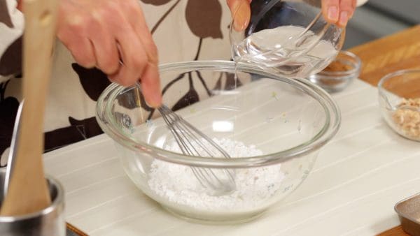 And now, let's make the mochi. In a microwave-safe bowl, place the shiratamako, sticky rice flour and add the water a little at a time while stirring with a balloon whisk. Be sure to mix thoroughly to avoid any pockets of dry flour.