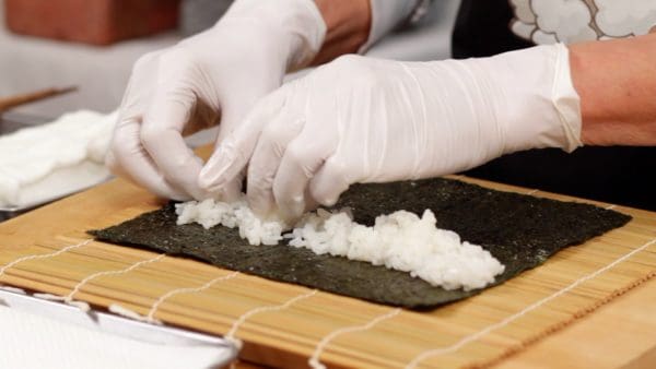 Mulai dari jarak sekitar 2 cm dari bagian atas rumput laut (nori), taruh satu porsi nasi sushi dan sebarkan secara horizontal dengan tangan.