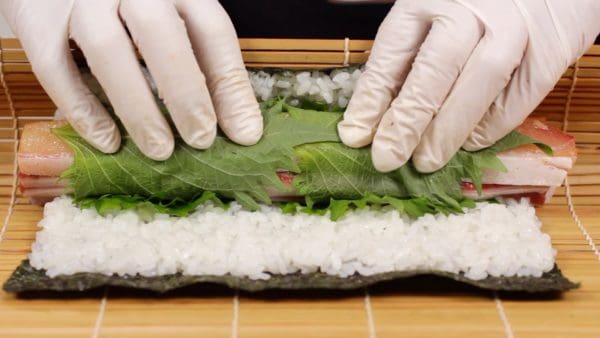 Para enrollar el sushi, levanta el borde inferior de la esterilla con los pulgares y sostén el pescado en su lugar con los otros cuatro dedos.