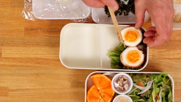 将一片红生菜叶放入便当盒中，将肉包好的鸡蛋放在生菜上，切面朝上。
