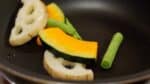 First, let's sauté the vegetables. In a frying pan, place the kabocha squash, lotus root, and string bean pods. Add a small amount of olive oil and toss to coat.
